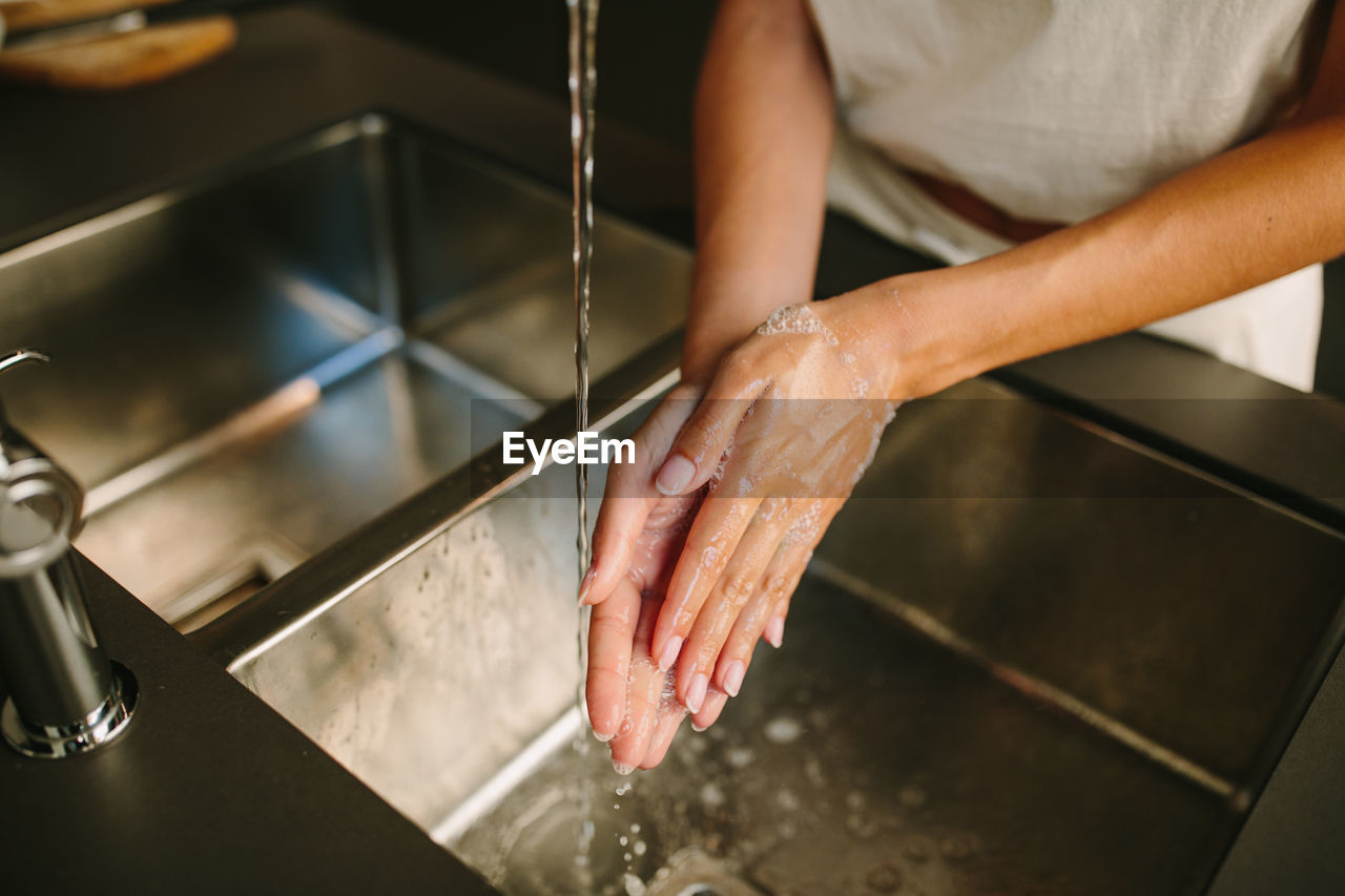 High angle of anonymous female washing hands with soap over metal sink in kitchen