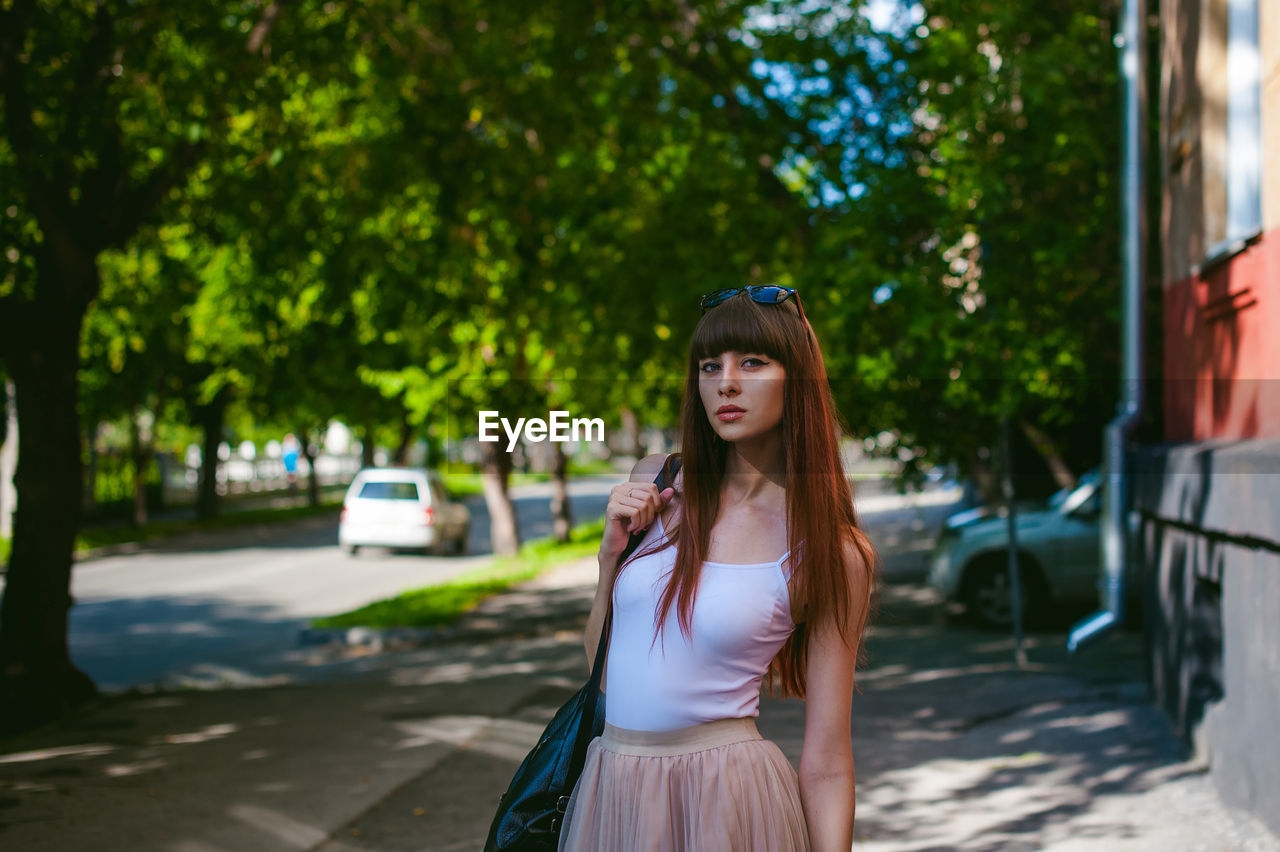 Portrait of young woman standing on sidewalk against trees