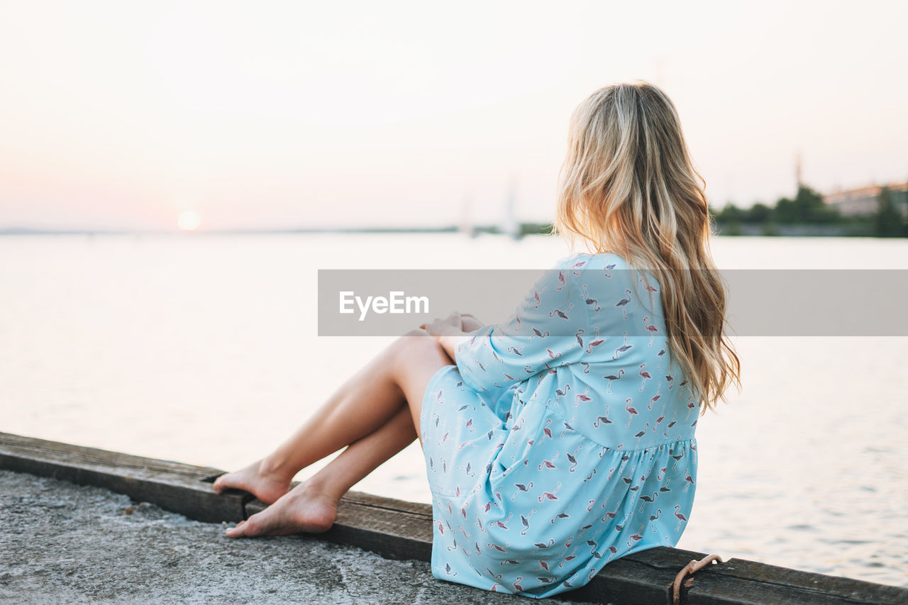 Beautiful blonde young woman in blue dress sitting on pier and looking on sunset