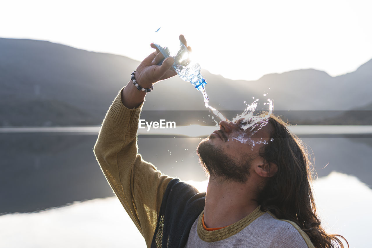 Man pouring water on face with bottle near lake during sunny day