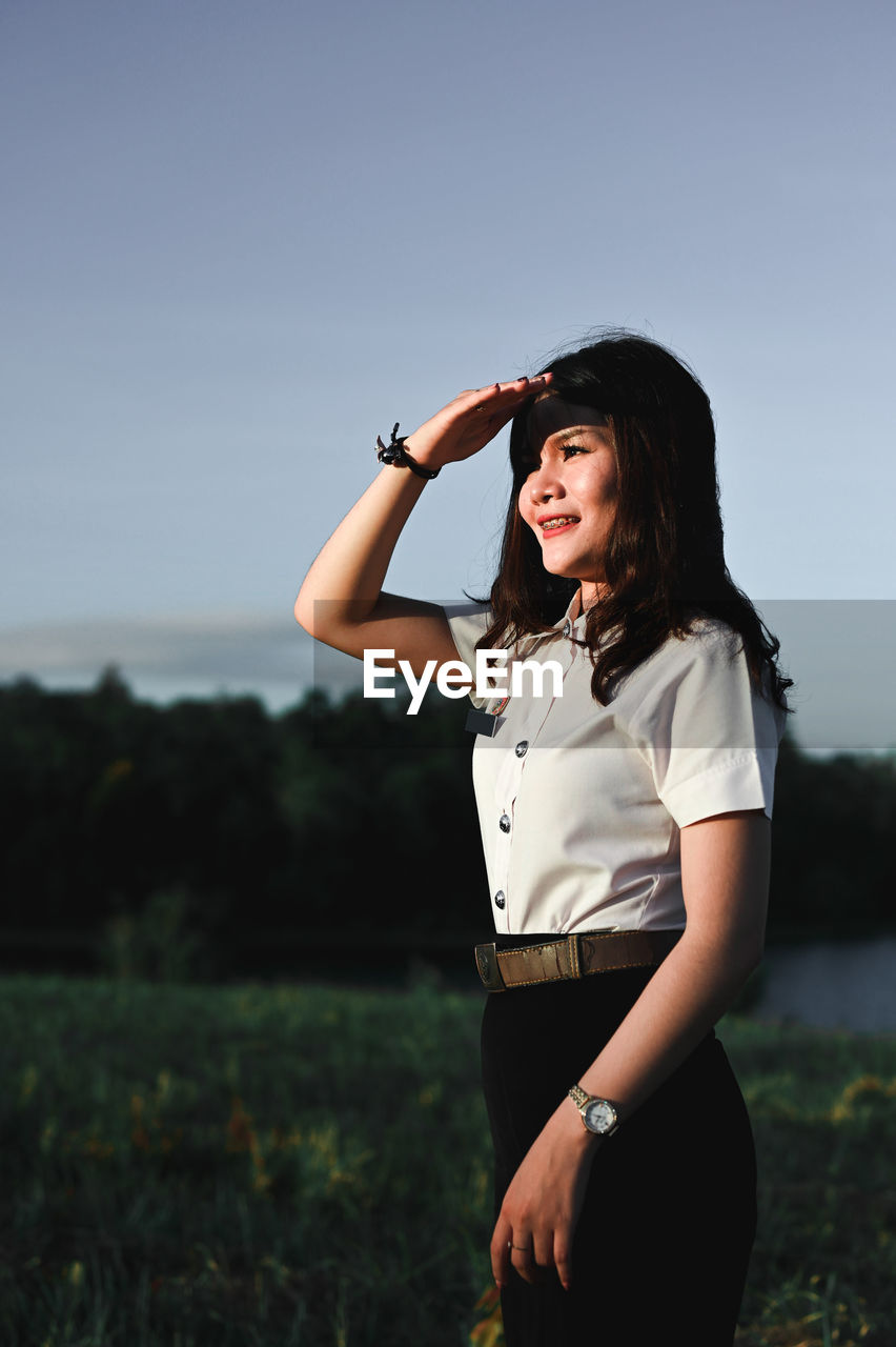 Smiling woman shielding eyes while standing on field against sky