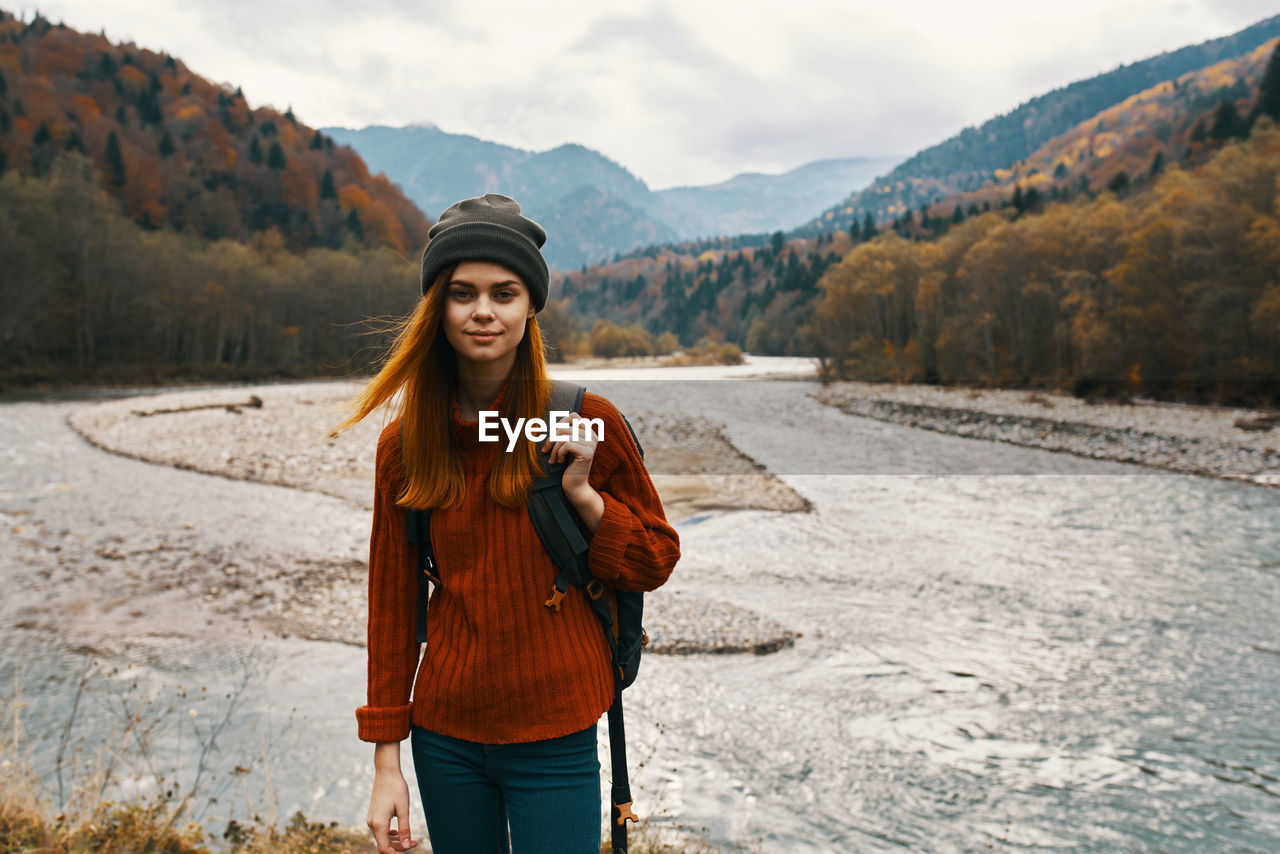 PORTRAIT OF SMILING YOUNG WOMAN STANDING AGAINST MOUNTAIN