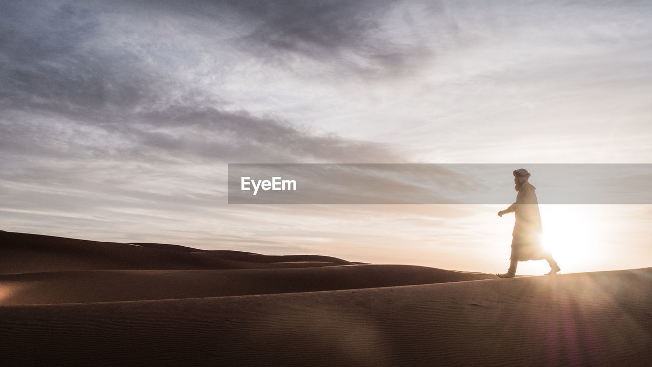 Man walking at desert against sky during sunset
