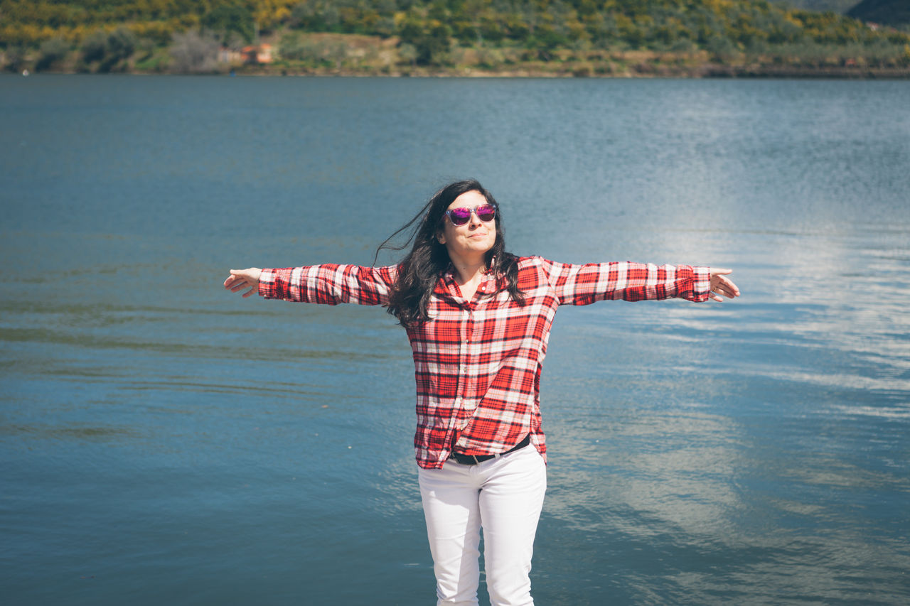 Happy woman with arms outstretched standing by lake during sunny day