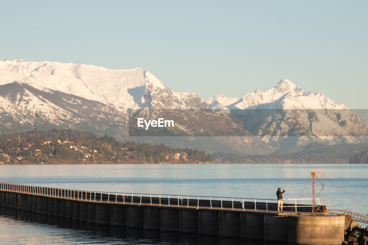 Man standing on pier over sea against snowcapped mountains