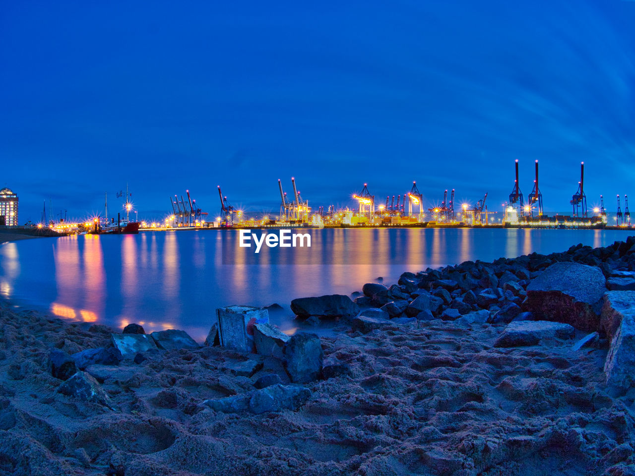 Illuminated factory by sea against sky at night