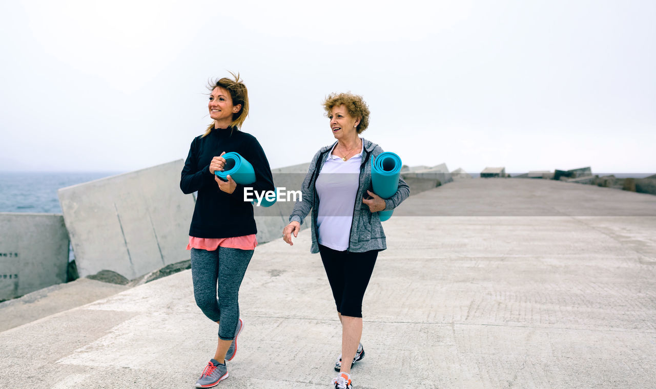 Full length of women with exercise mats by sea on walkway