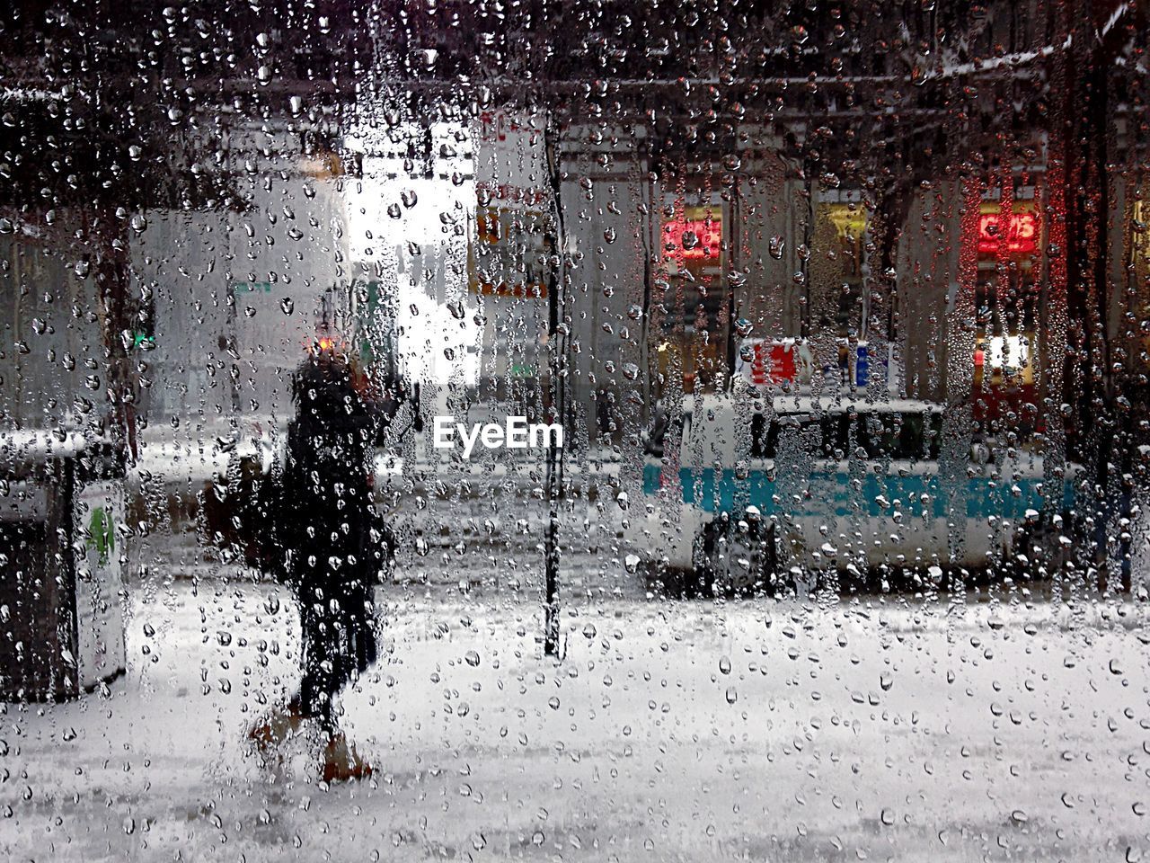 Woman walking on sidewalk during rainy season seen through wet glass window