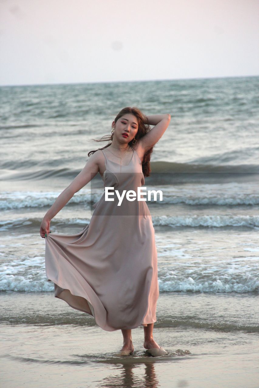 Full length of mature woman wearing dress while posing at beach against sky