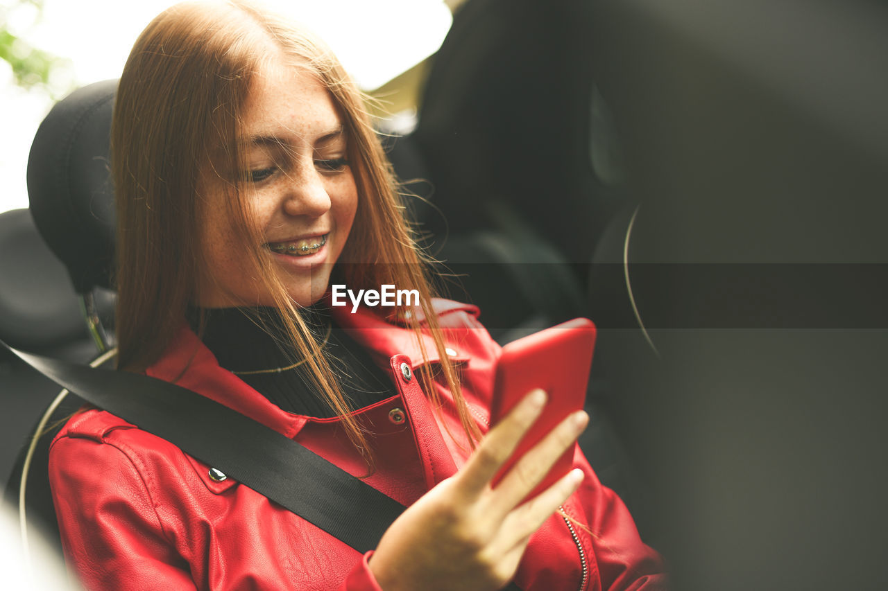 Trendy girl sitting in a car with security belt. smiling teen using smartphone and chat with friend.