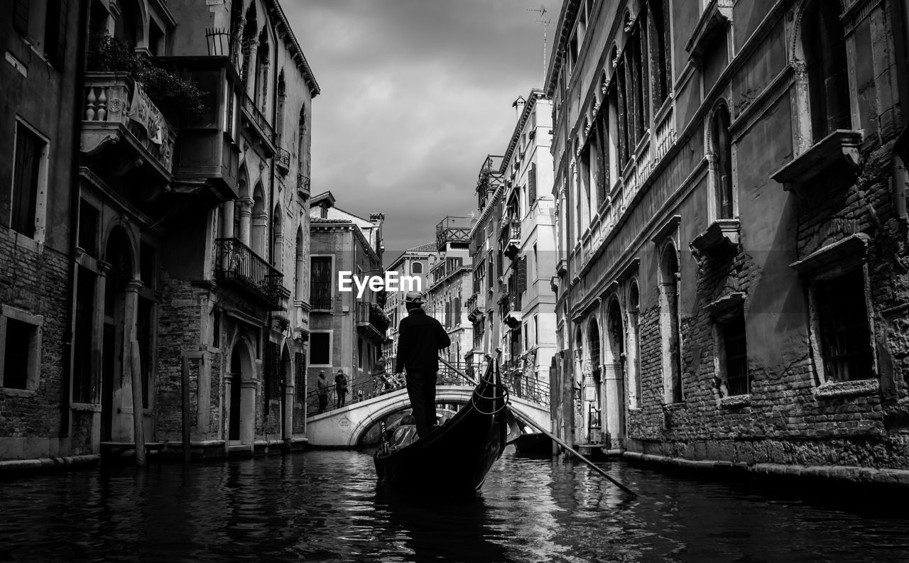 Rear view of gondolier sailing boat in canal amidst buildings