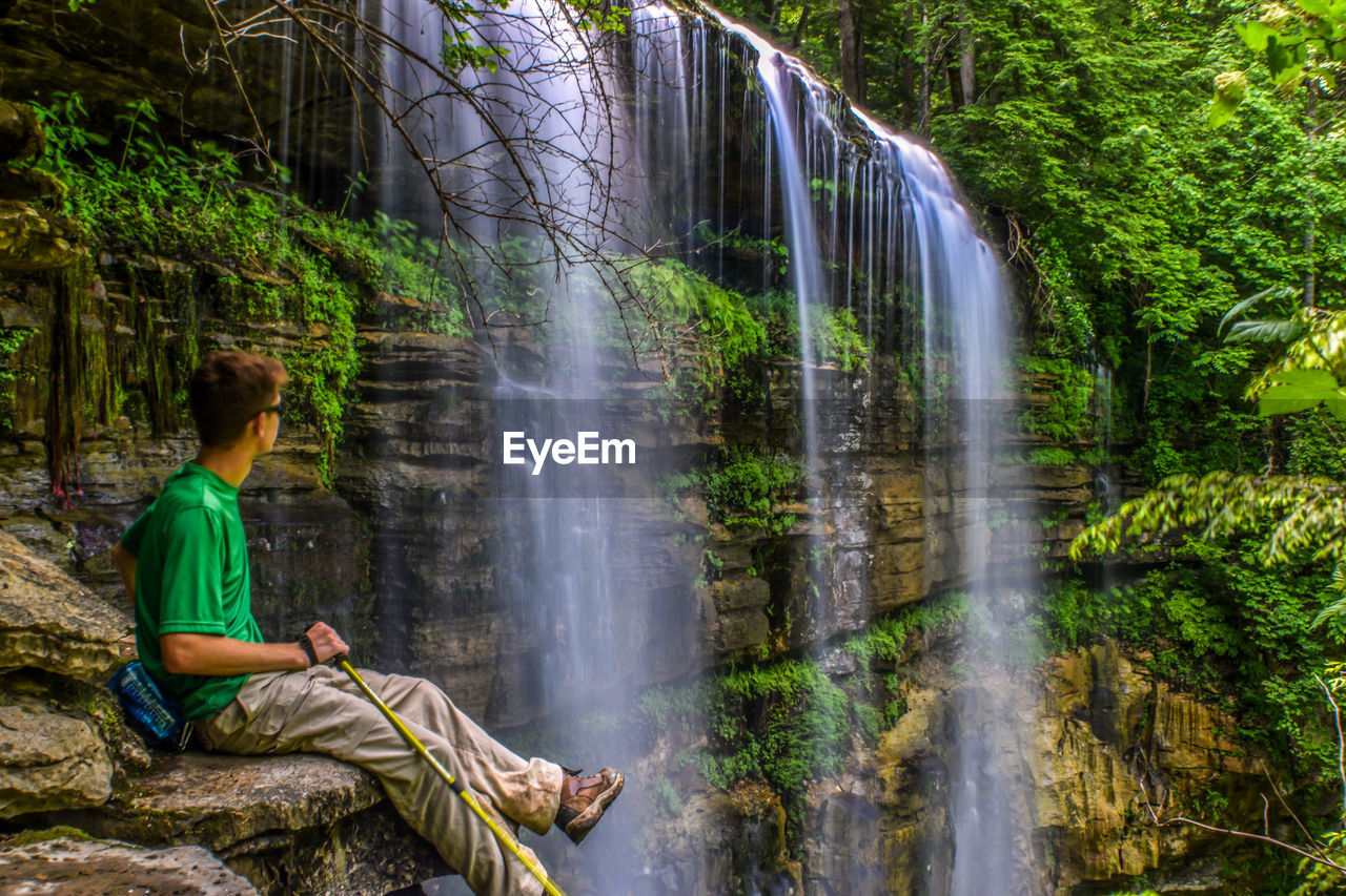 Young man sitting on rock while looking at waterfall