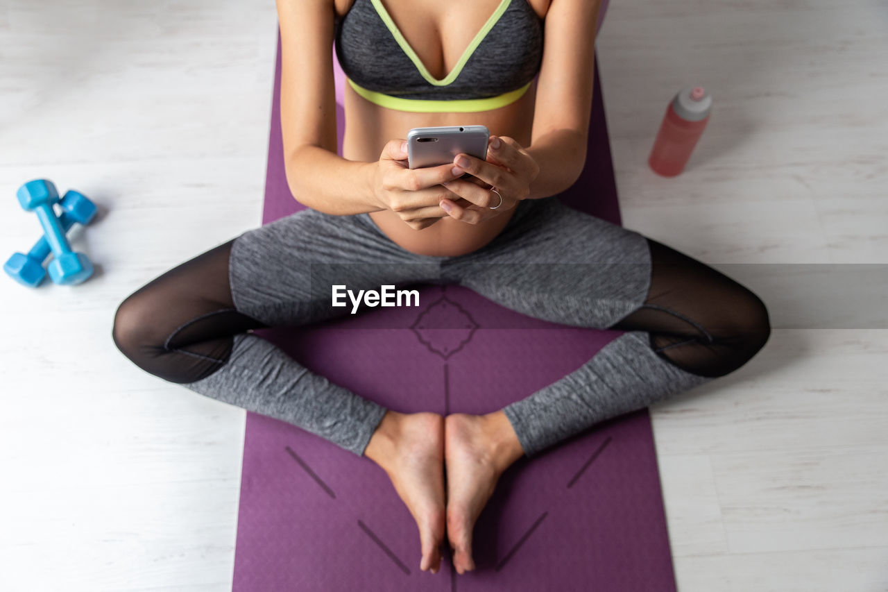 From above of pregnant female in sportswear sitting on mat resting after in yoga practice using mobile phone