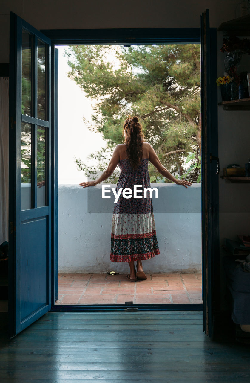 Full body back view of unrecognizable female in casual sundress admiring view from balcony of country house while spending summer holidays in tropical countryside