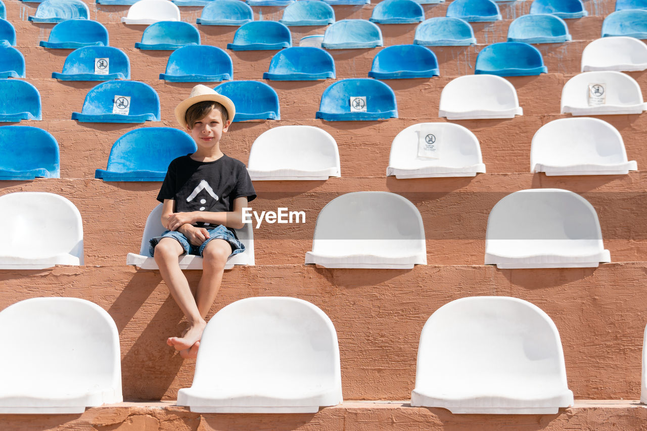 Serious boy in summer clothes sitting on one of chair on stadium and looking to camera