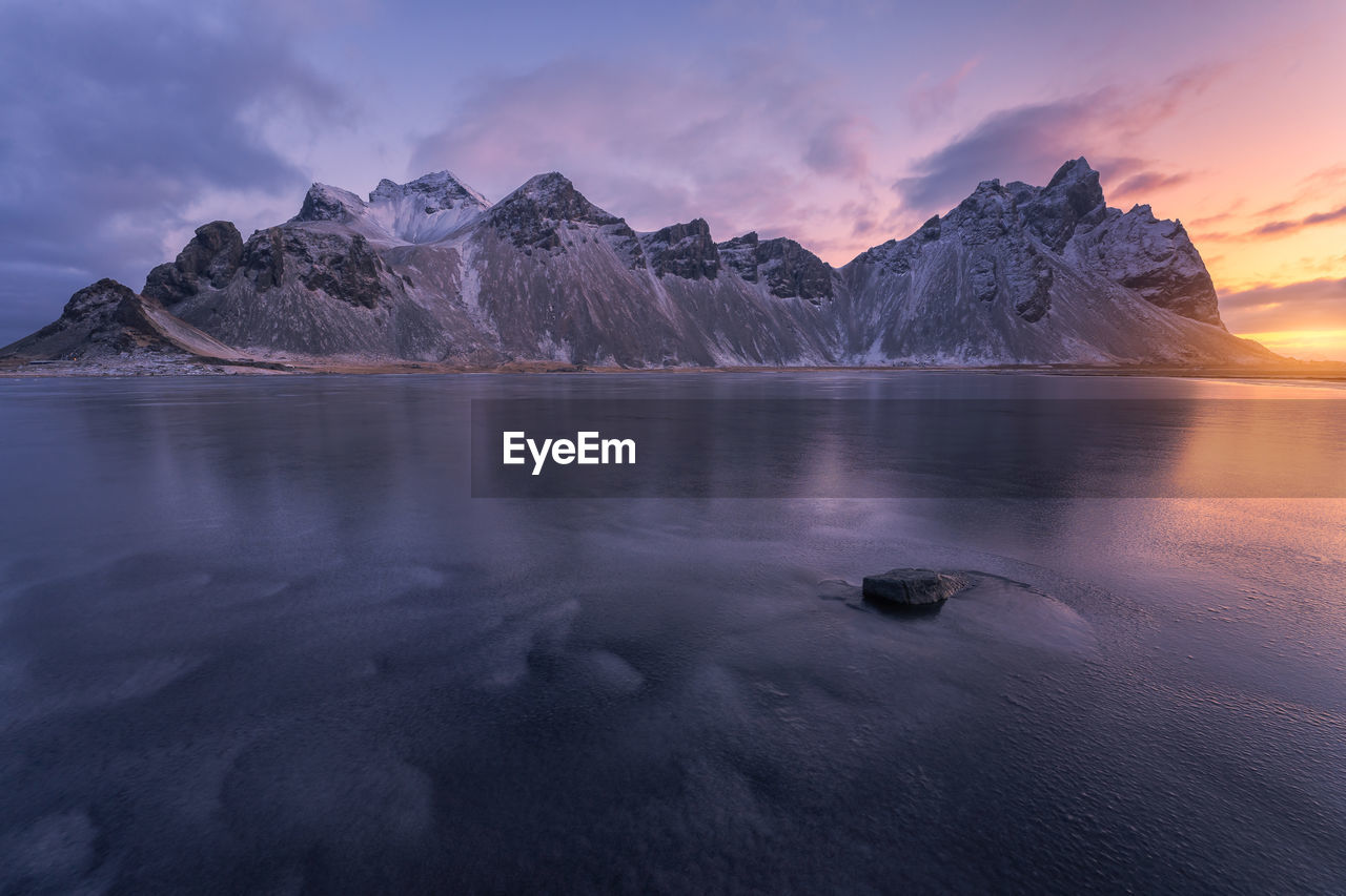 Spectacular nordic scenery of calm frozen lake near rocky vestrahorn mountain with snowy peaks during colorful sunset at stockness beach, iceland