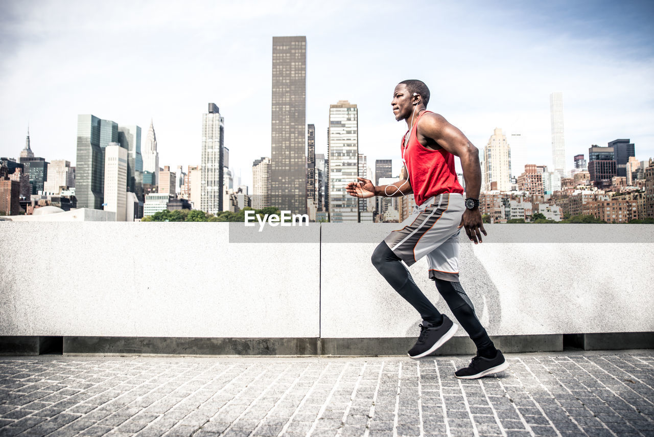 Full length of man running while listening music against buildings and sky in city