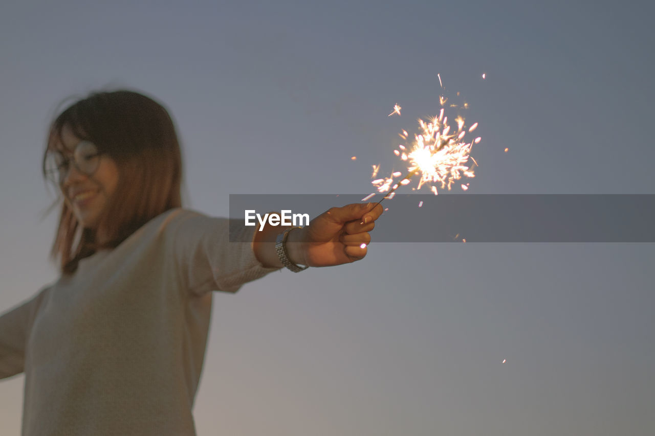 LOW ANGLE VIEW OF HAND HOLDING SPARKLER AGAINST CLEAR SKY