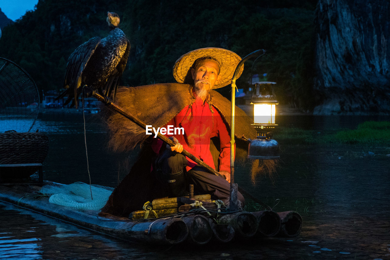 Portrait of mature fisherman with cormorant by illuminated oil lantern on wooden raft in river