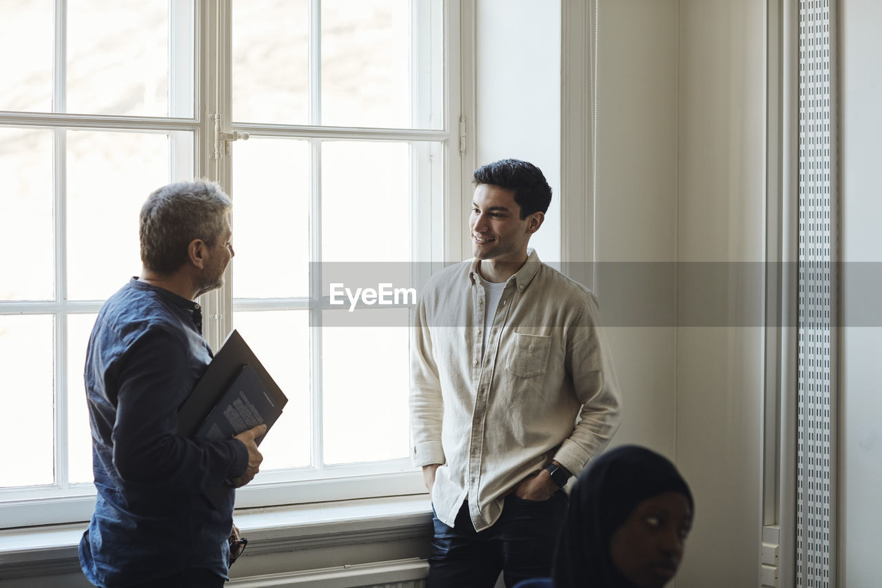 Male teacher talking with student while standing by window in university