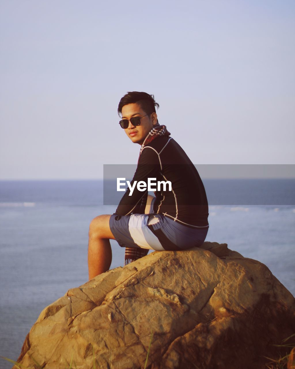 Portrait of young man in sunglasses sitting on rock at beach against clear sky