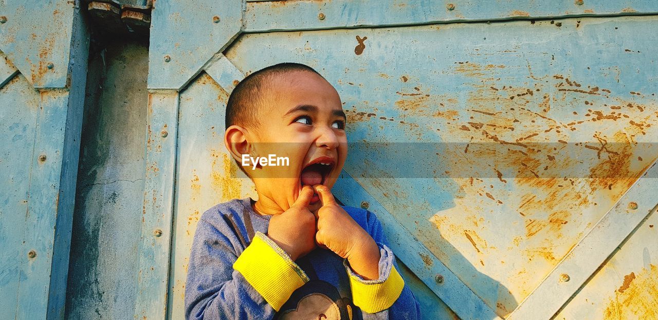 Cute boy with finger in mouth standing against metal gate