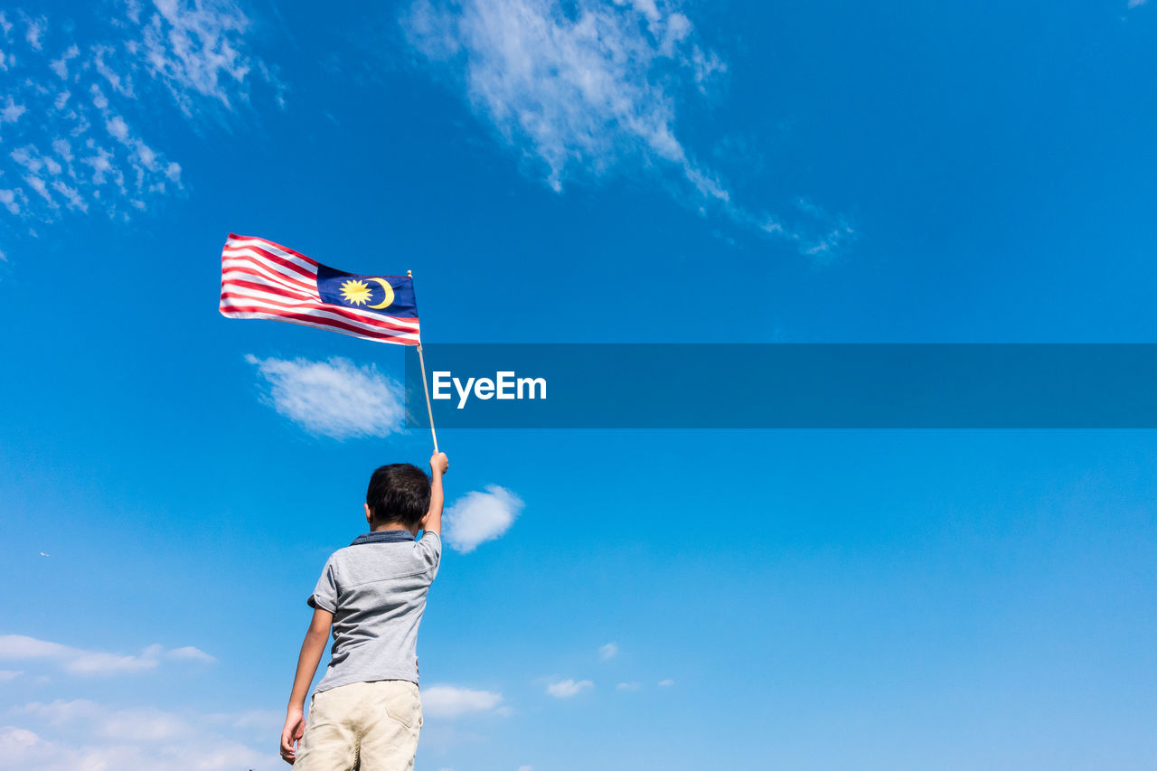 Rear view of boy holding malaysian flag while standing against blue sky