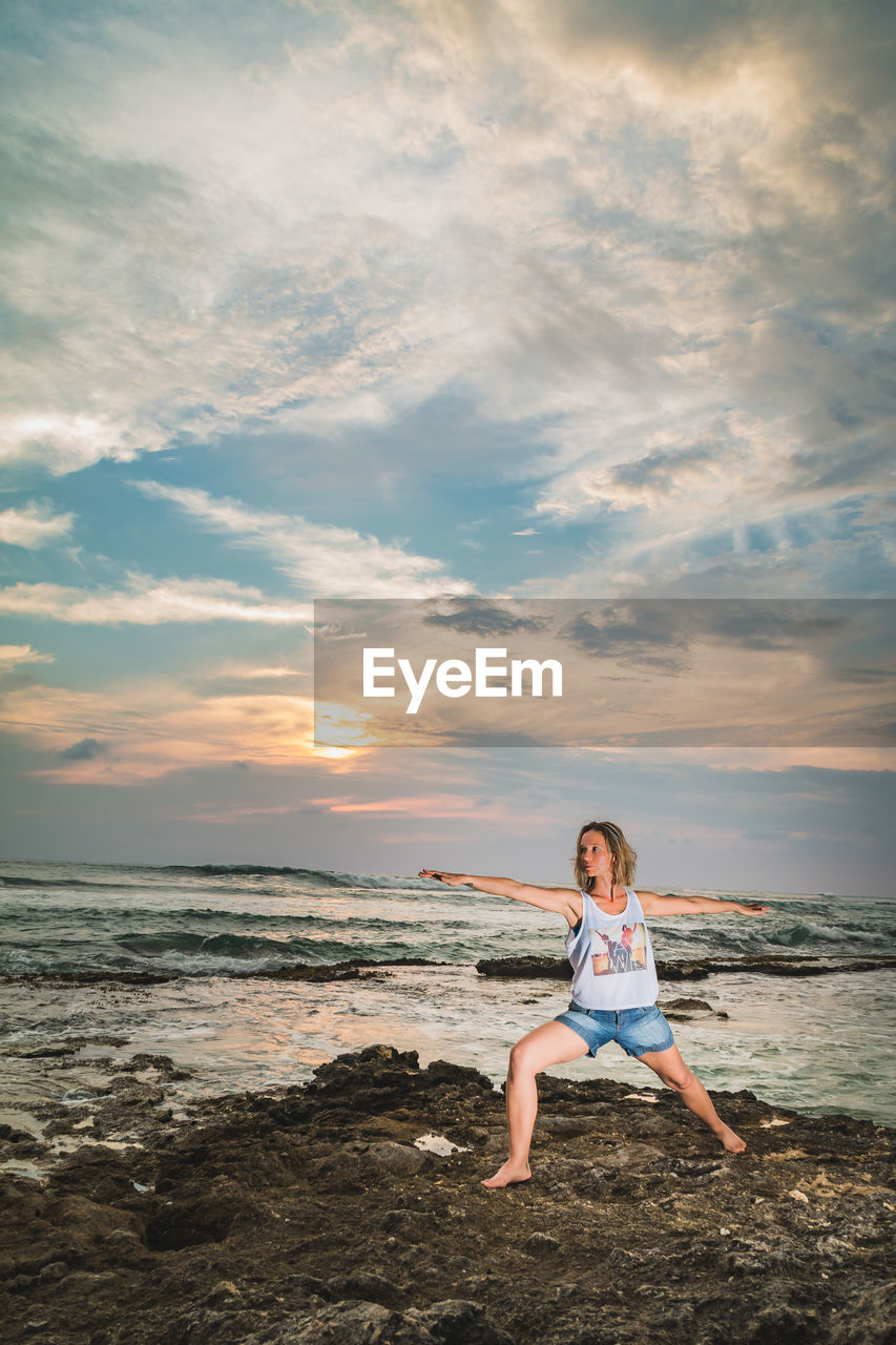 Woman practicing yoga on rocky shore against cloudy sky