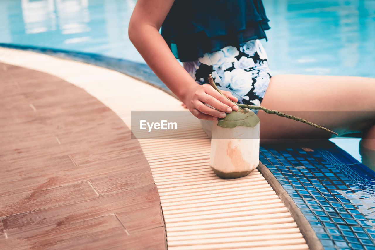 Midsection of woman sitting with coconut at poolside