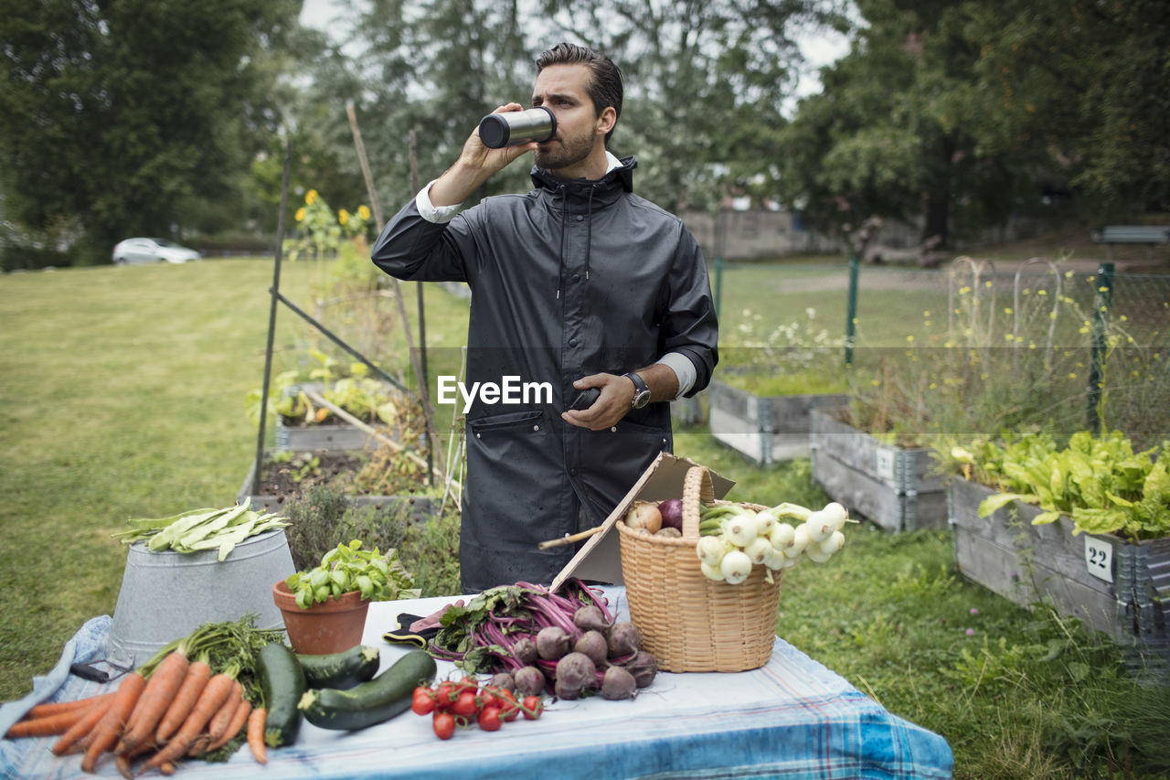 Mid adult man drinking coffee while standing by freshly harvested vegetables on table at urban garden