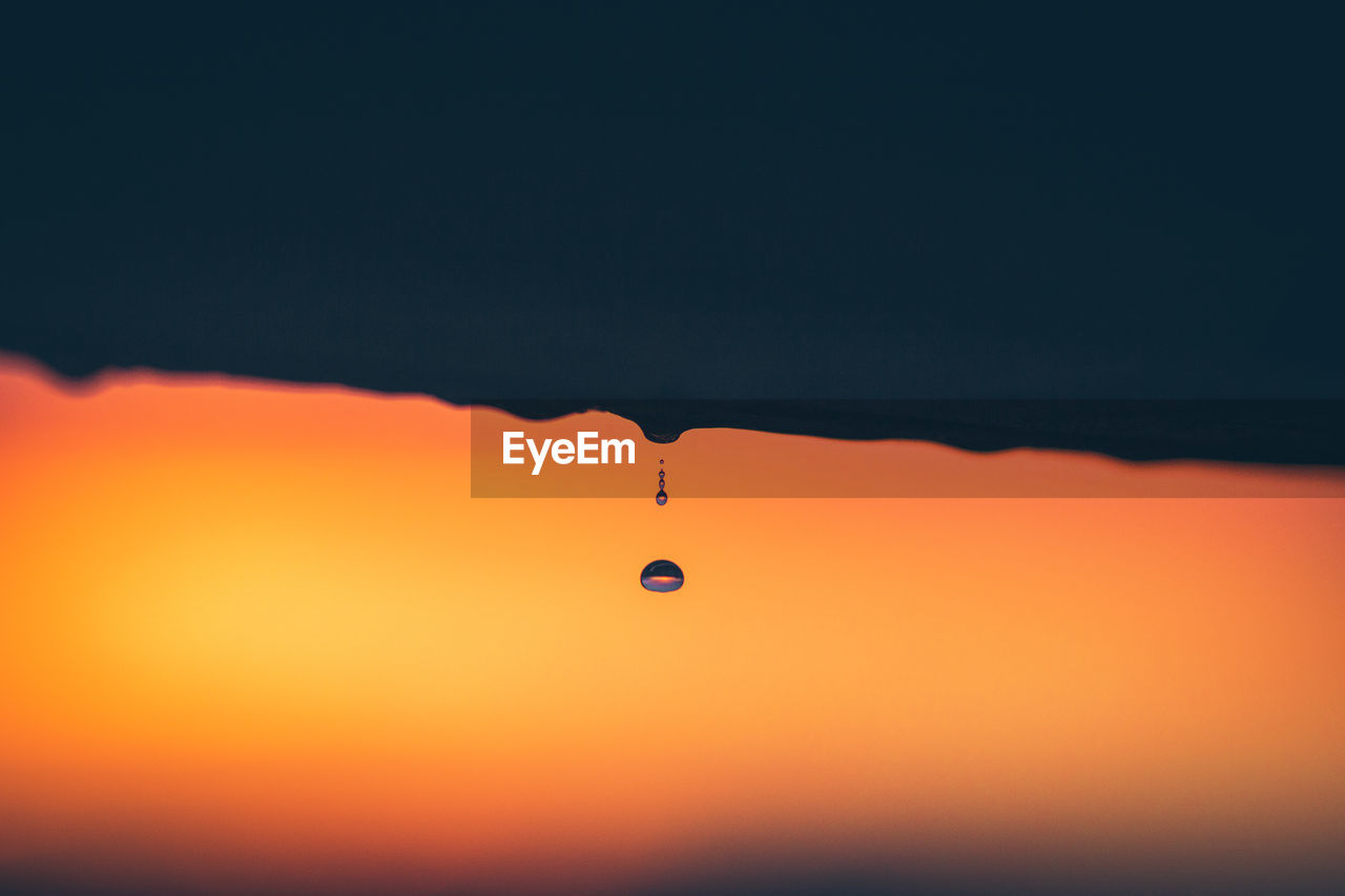 Close-up of drop falling from railing against sky during sunset