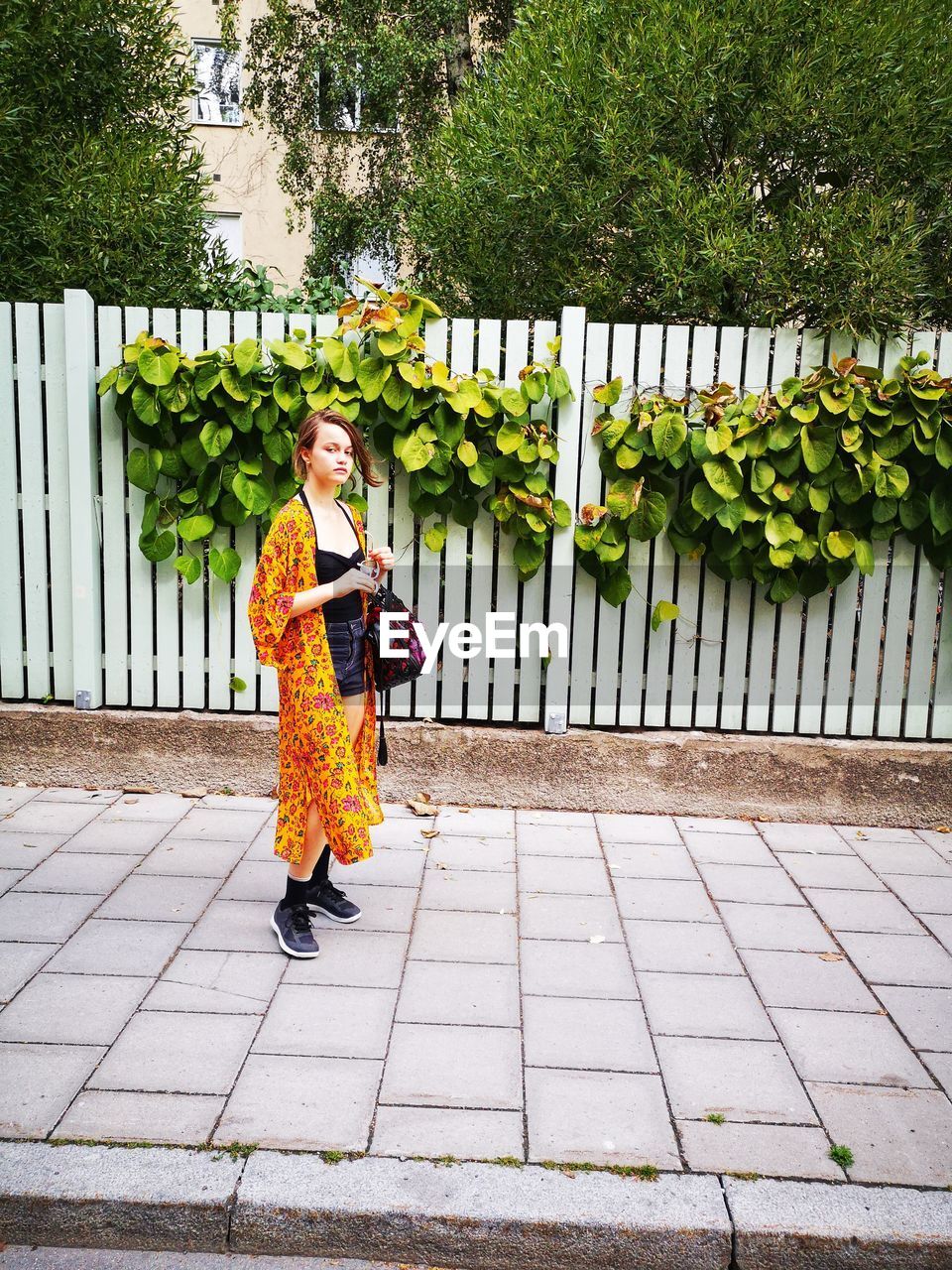 Full length portrait of girl standing on footpath against fence