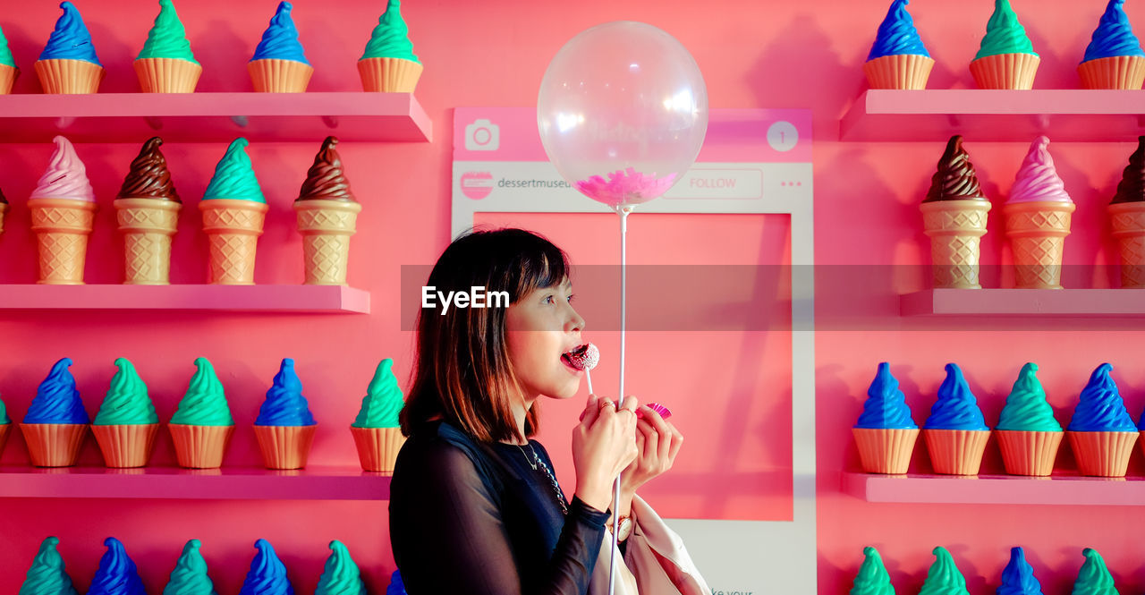 Woman holding balloon and eating cake pop while standing against wall