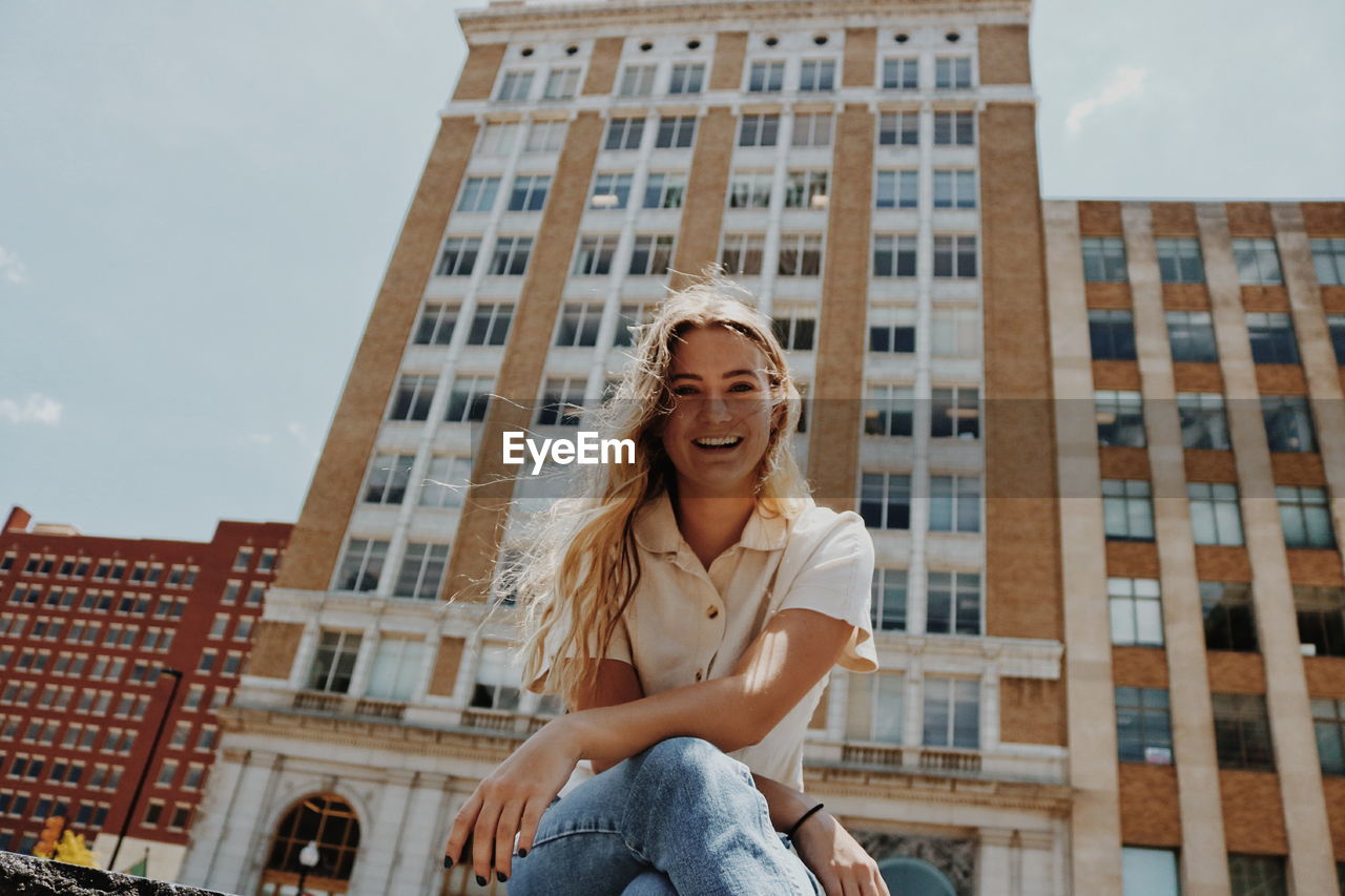 Low angle portrait of smiling young woman sitting against building in city