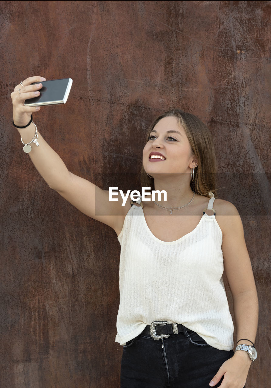 Smiling young woman taking selfie with mobile phone against wall