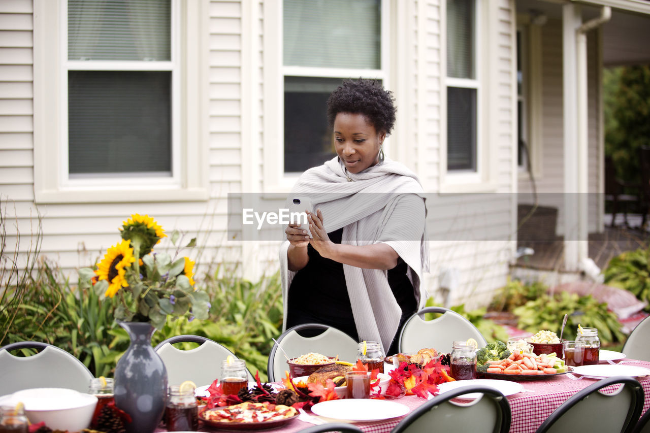 Woman photographing vase through phone at table against house