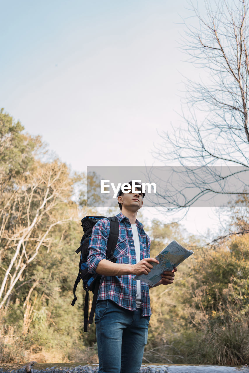 Low angle of male hiker with backpack navigating with paper map while standing in woods and looking away