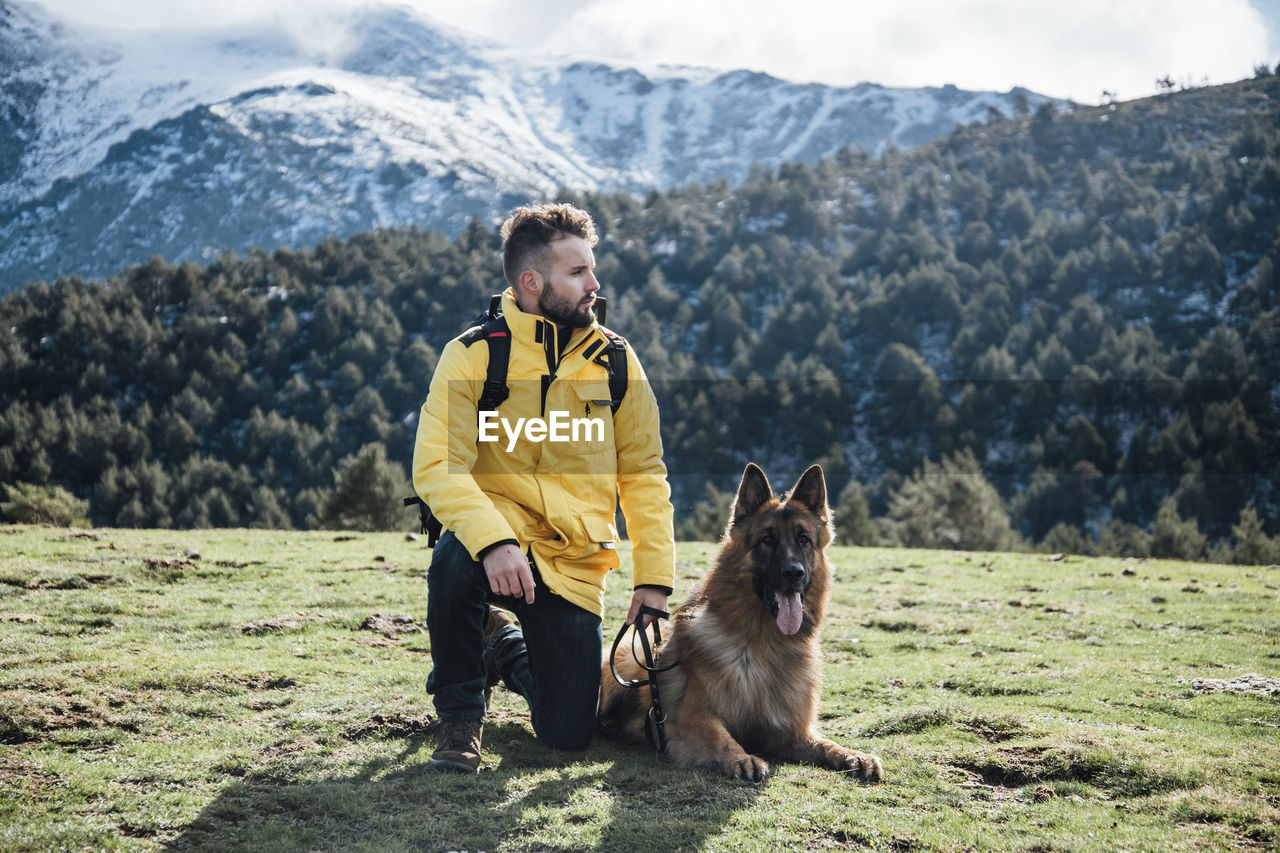 Young man with yellow jacket and backpack plays with german shepherd dog in the mountains.