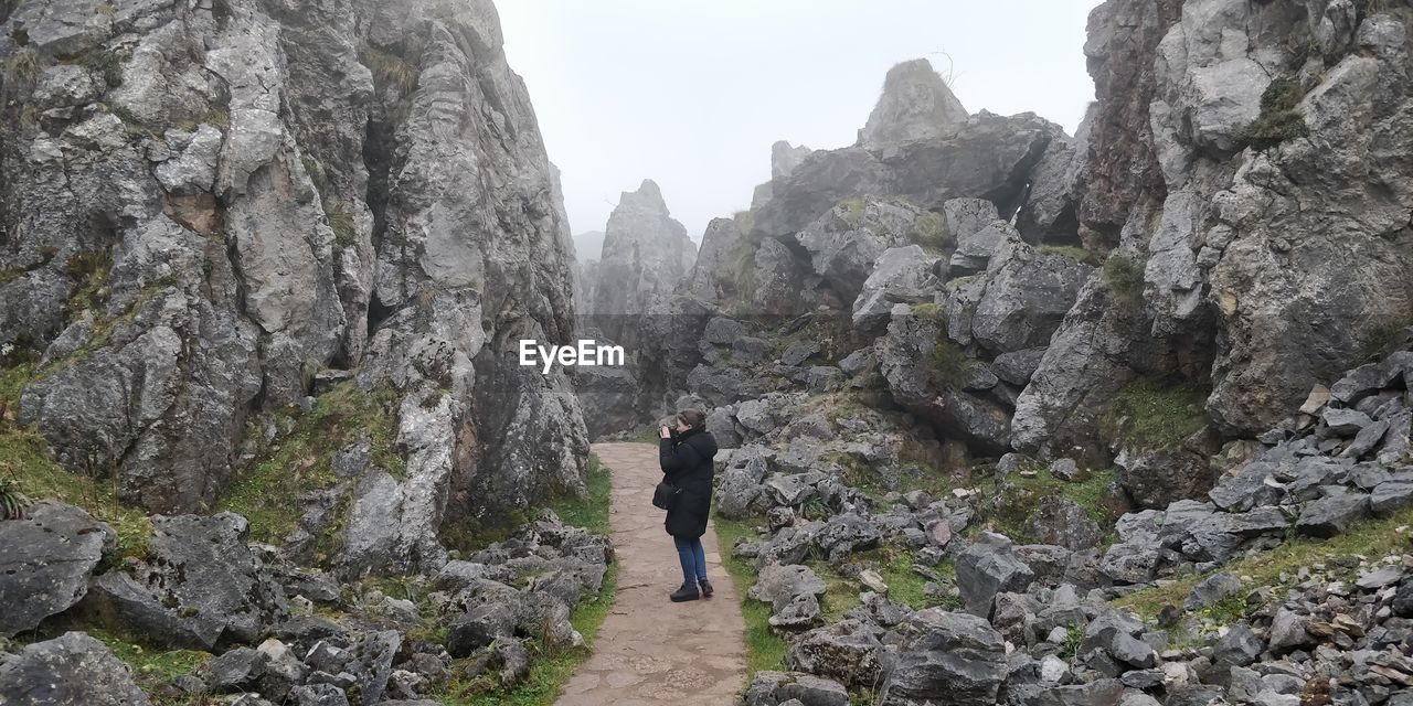 REAR VIEW OF MAN STANDING ON ROCKS AGAINST MOUNTAINS