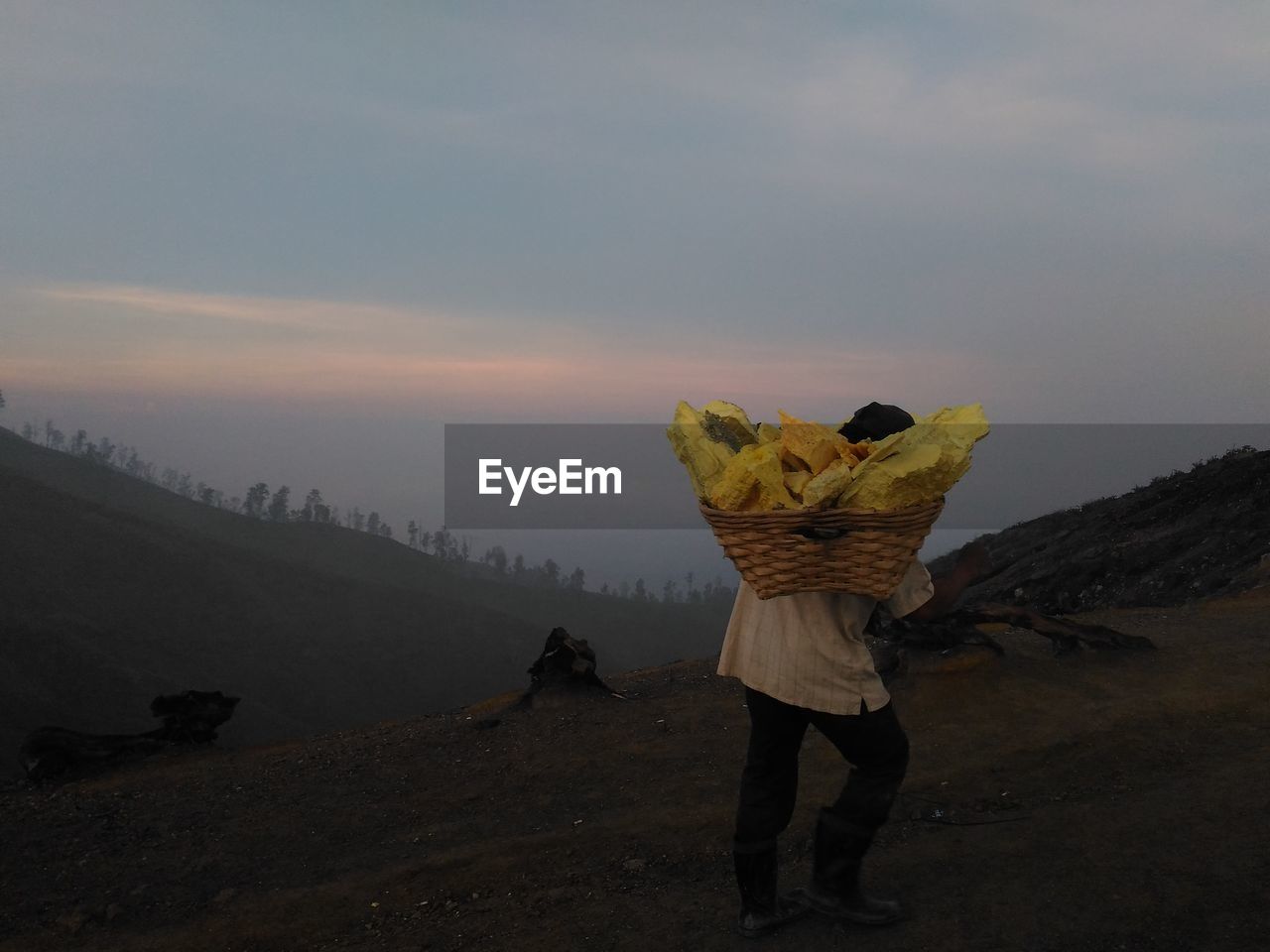 Rear view of worker carrying yellow stones in basket during sunset