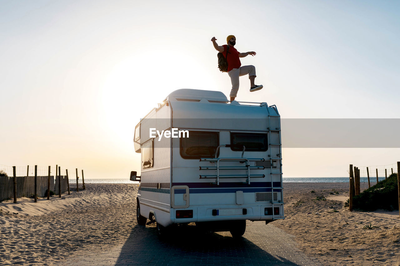 Cheerful male explorer standing on roof of camper parked on seashore and enjoying with outstretched arms and raised leg against sundown sky