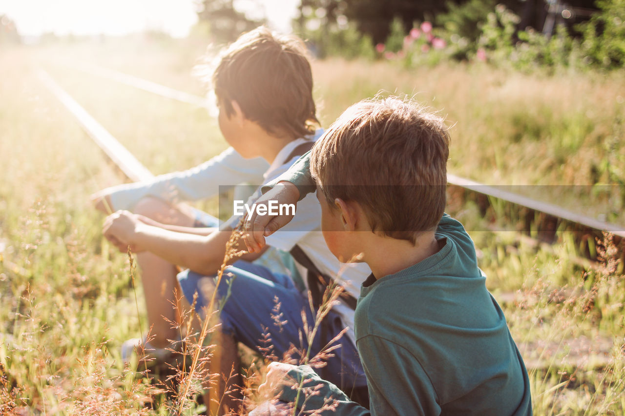 Boys sitting on railroad track during sunny day