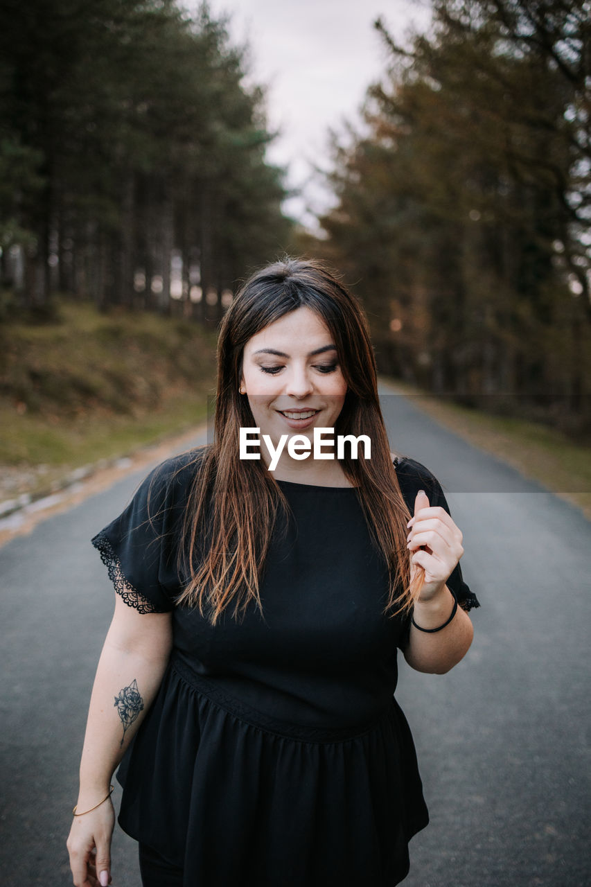 Content female standing on empty roadway surrounded by green coniferous woods and looking down