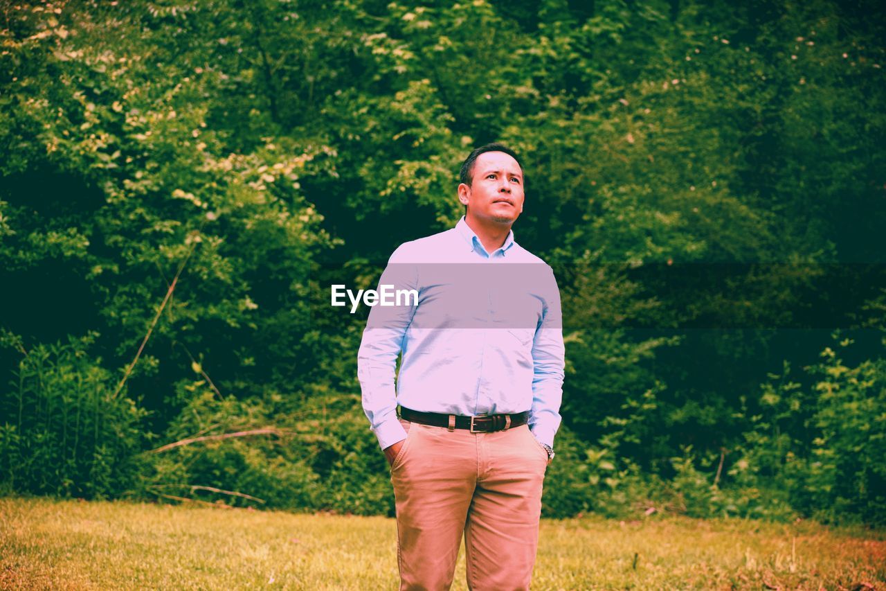 Thoughtful man with hands in pockets standing on grassy field against trees