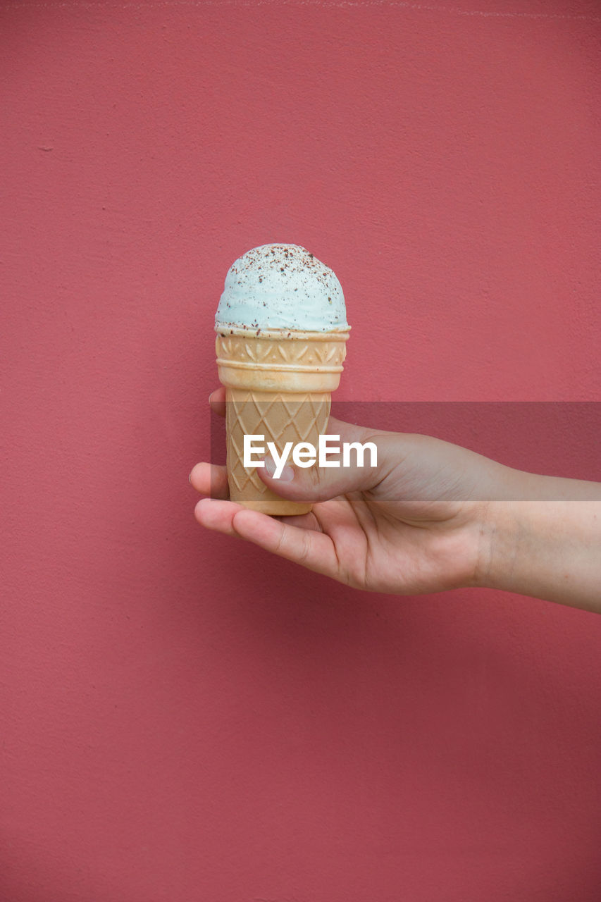 HUMAN HAND HOLDING ICE CREAM CONE AGAINST RED WALL