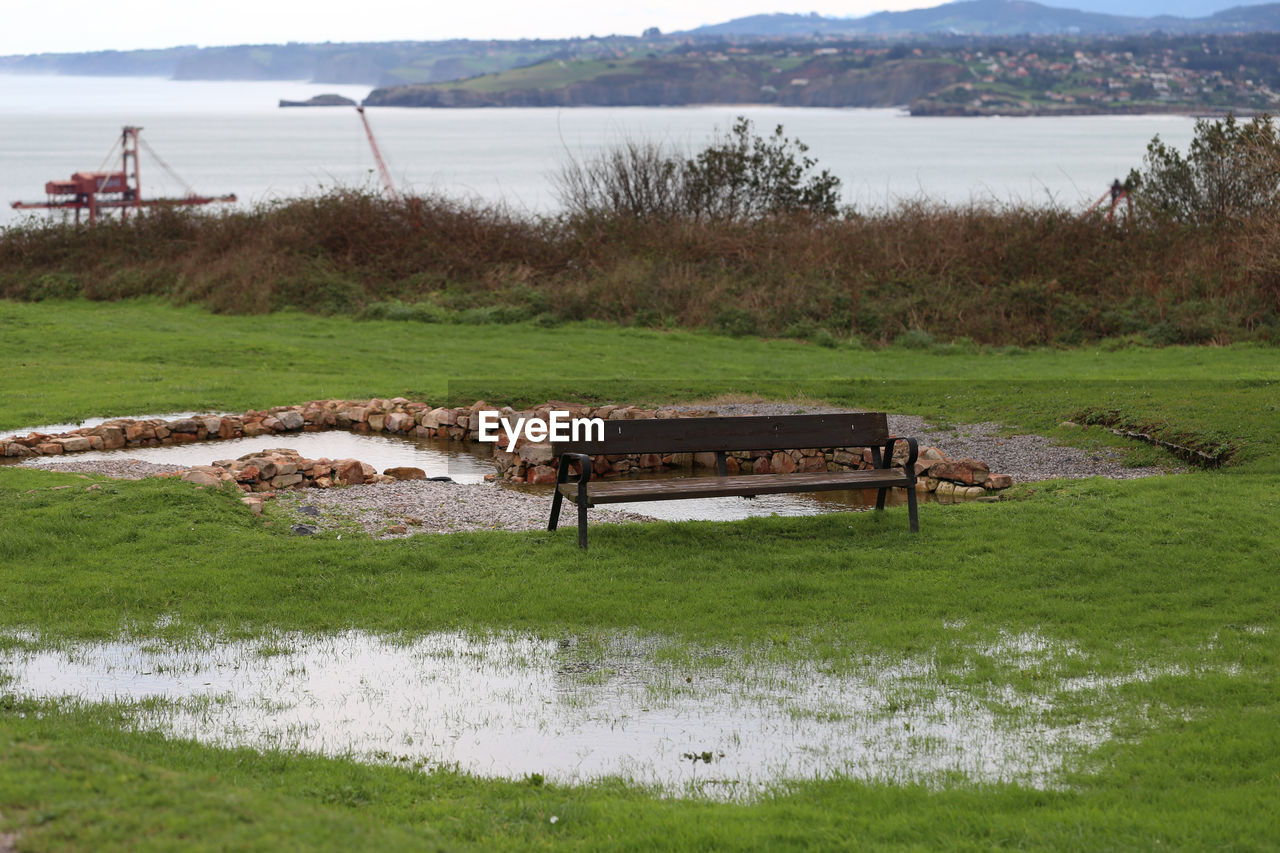 EMPTY BENCH ON FIELD BY LAKE AGAINST SKY