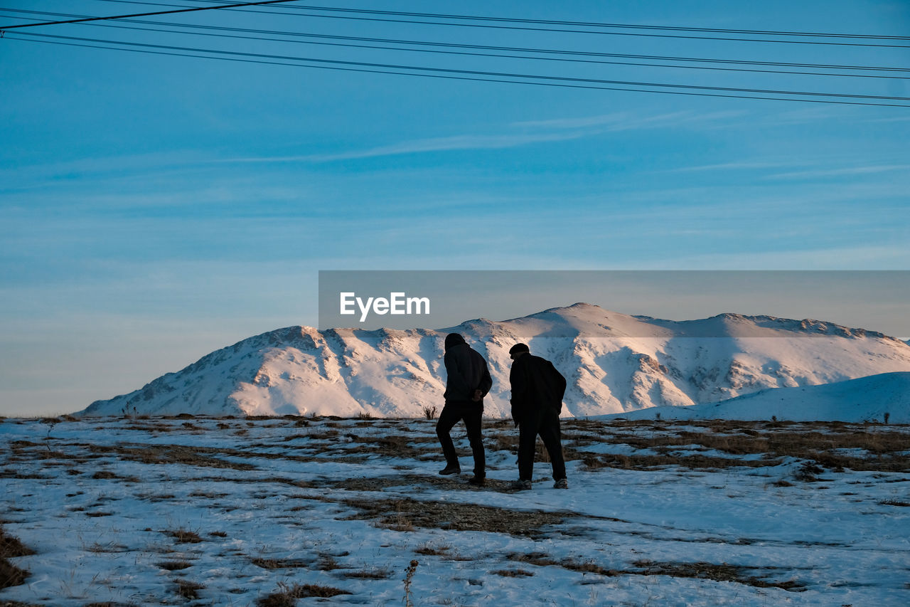 REAR VIEW OF MEN WALKING ON SNOWCAPPED MOUNTAINS AGAINST SKY