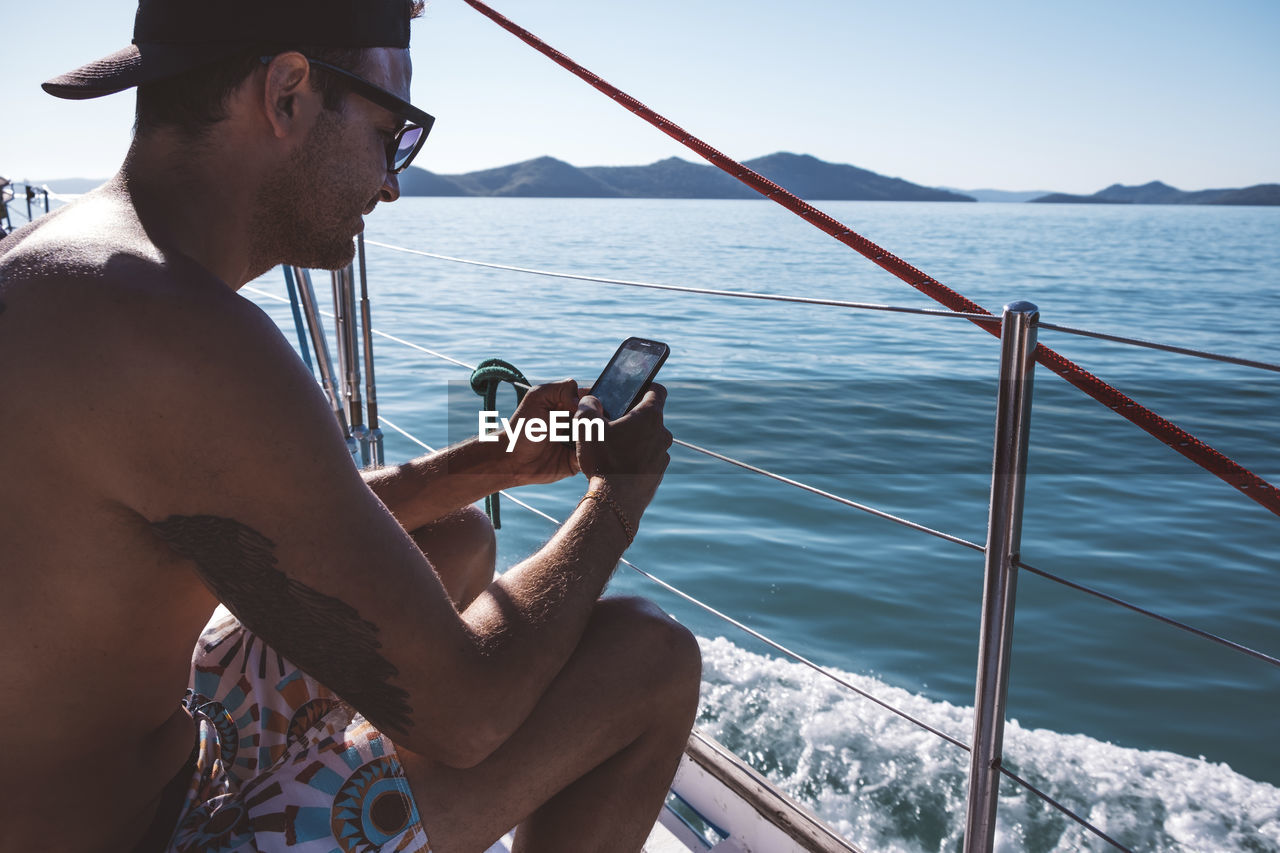 Side view of shirtless man using smart phone while sitting in boat on sea against clear sky during sunny day
