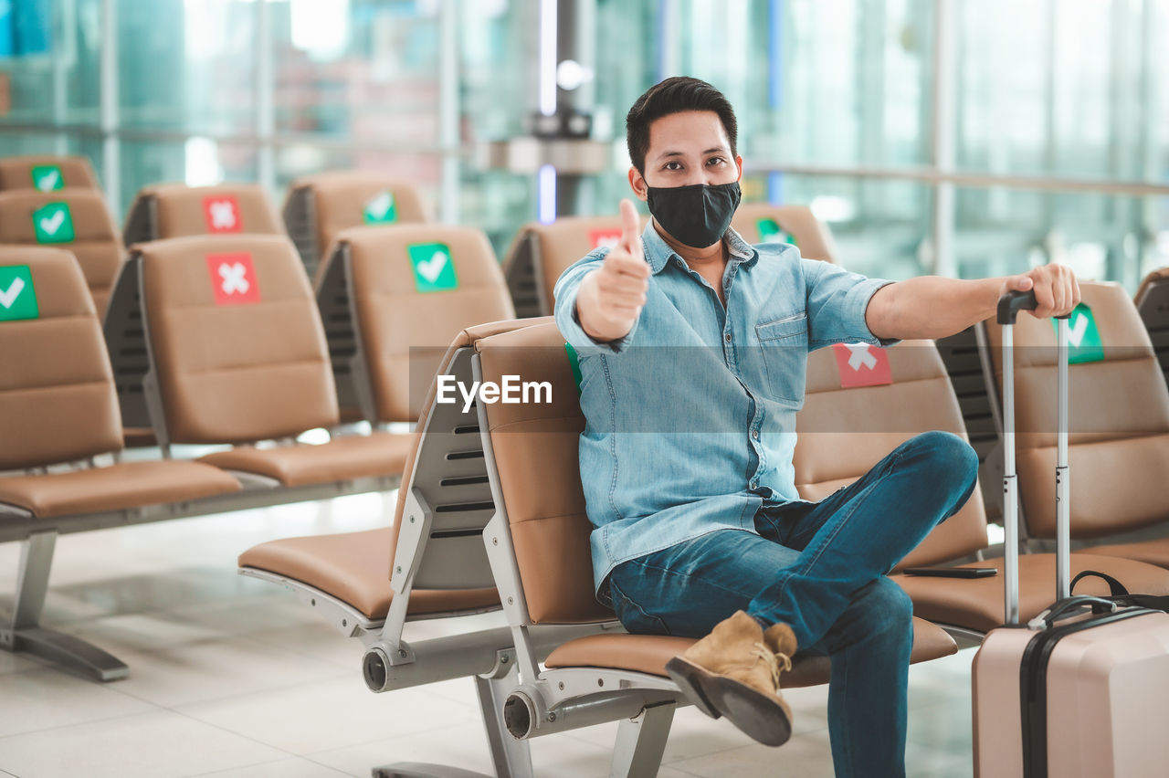 Portrait of mid adult man wearing flu mask gesturing while sitting at airport