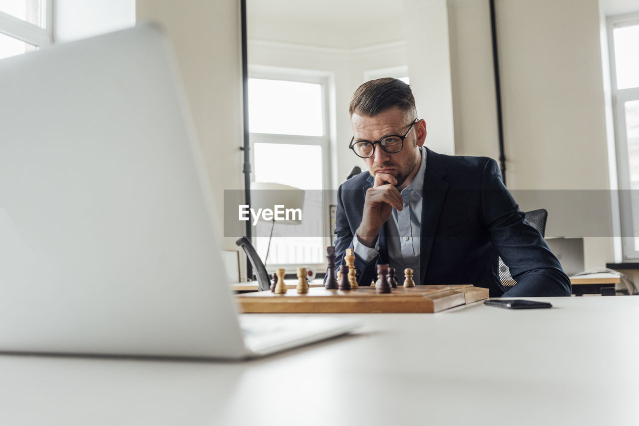 Businessman looking at laptop while playing chess at office